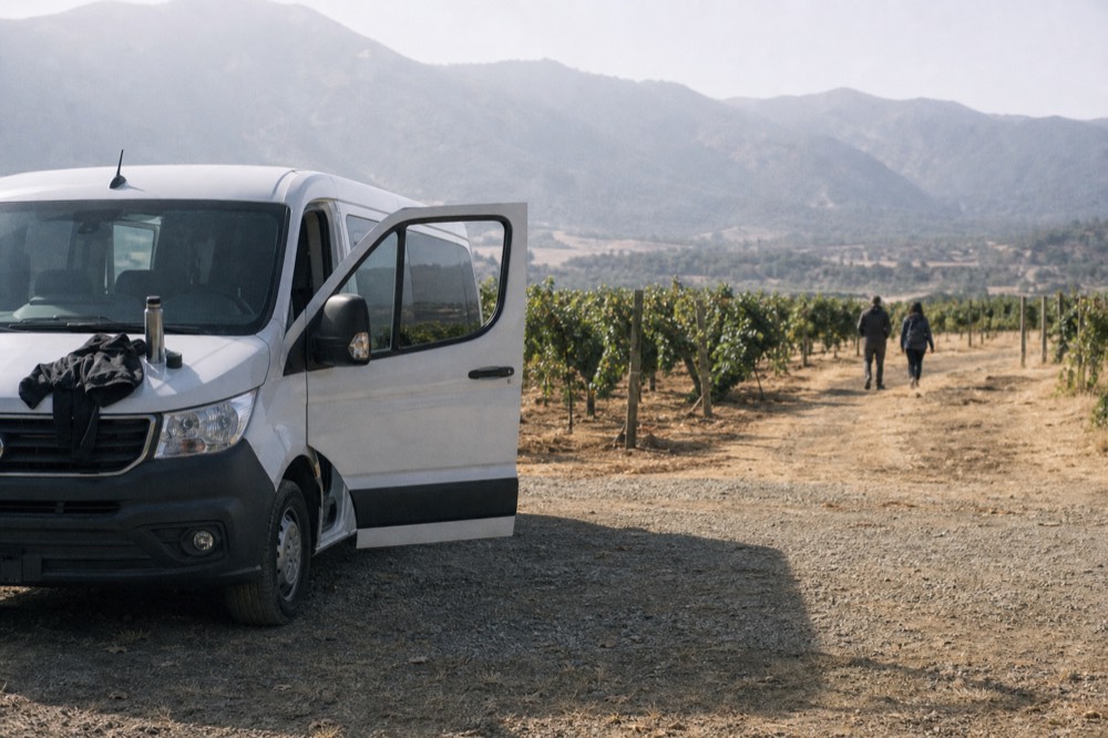 A driver resting against a tour van parked on a gravel road beside vineyard rows, no text, no logos, no people